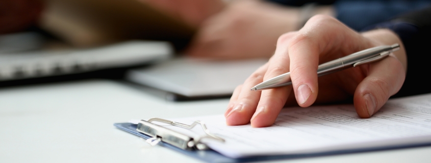 Hand of businessman in suit filling and signing with silver pen partnership agreement form clipped to pad closeup. Management training course, some important document, team leader ambition concept