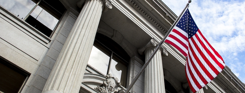 Stone granite official government building in city with American flag waving and flying in the wind, looking up, outside, judicial, freedom, civil courts, columns, criminal justice, law